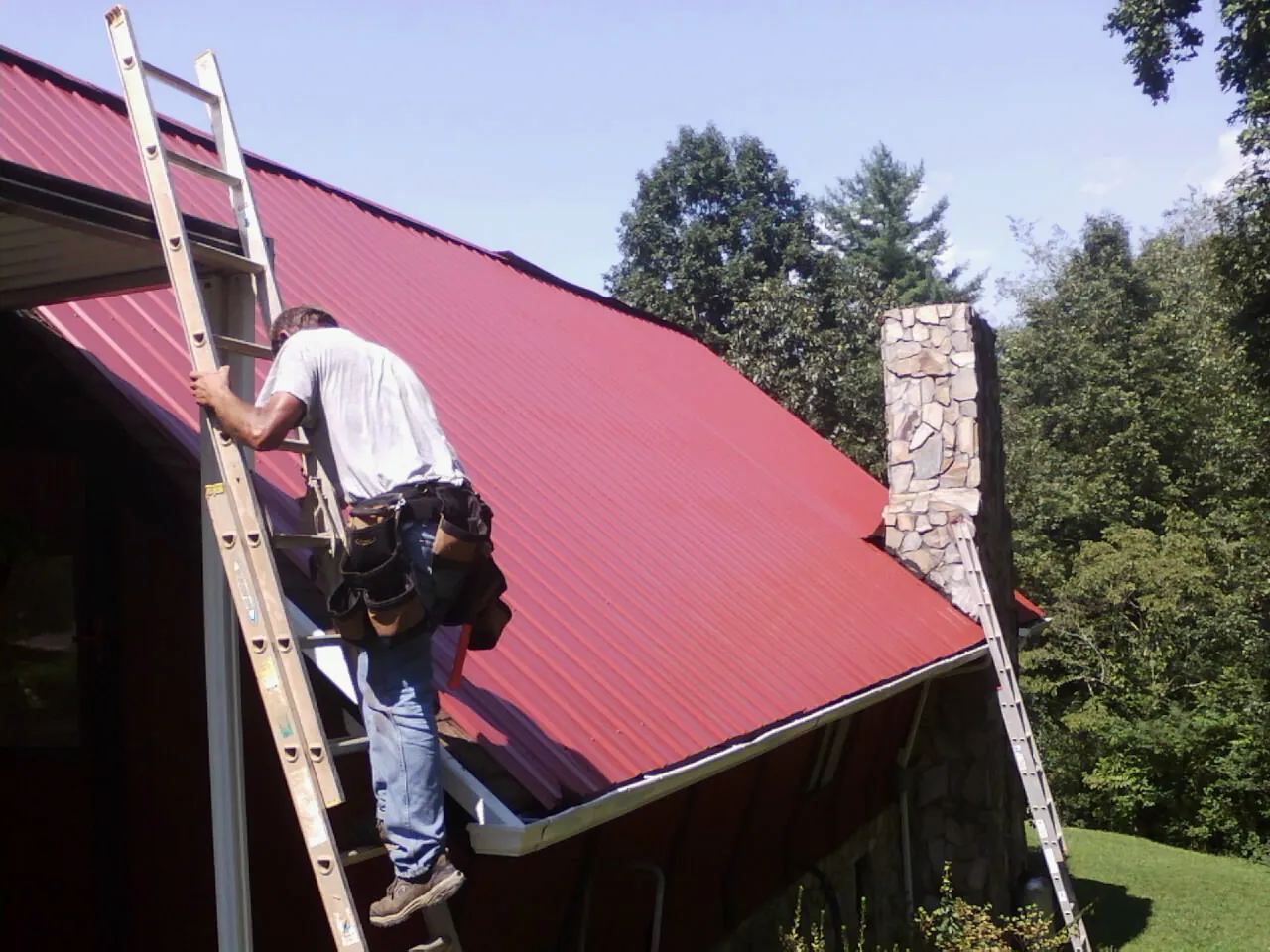 Roofer on a ladder beside a red metal roof and stone chimney — Soffit & Fascia Repair by Roofing Brothers in Saint Pauls